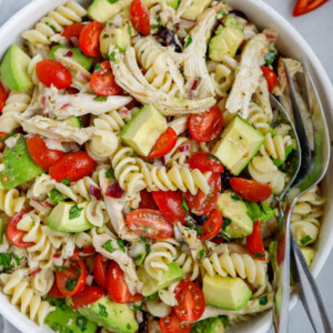 A bowl of avocado chicken pasta salad with a fork and spoon in the bowl.