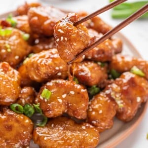 A plate with Crispy Honey Chicken with a pair of chopsticks lifting a piece up.