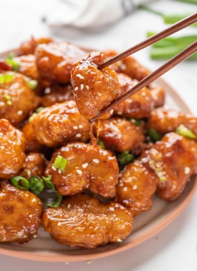 A plate with Crispy Honey Chicken with a pair of chopsticks lifting a piece up.