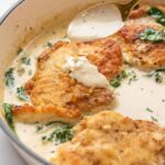 Golden-brown chicken breasts with wilted spinach in a creamy skillet sauce as a spoon pours more sauce over the top.