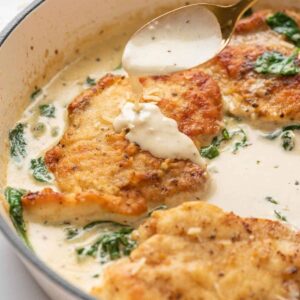 Golden-brown chicken breasts with wilted spinach in a creamy skillet sauce as a spoon pours more sauce over the top.