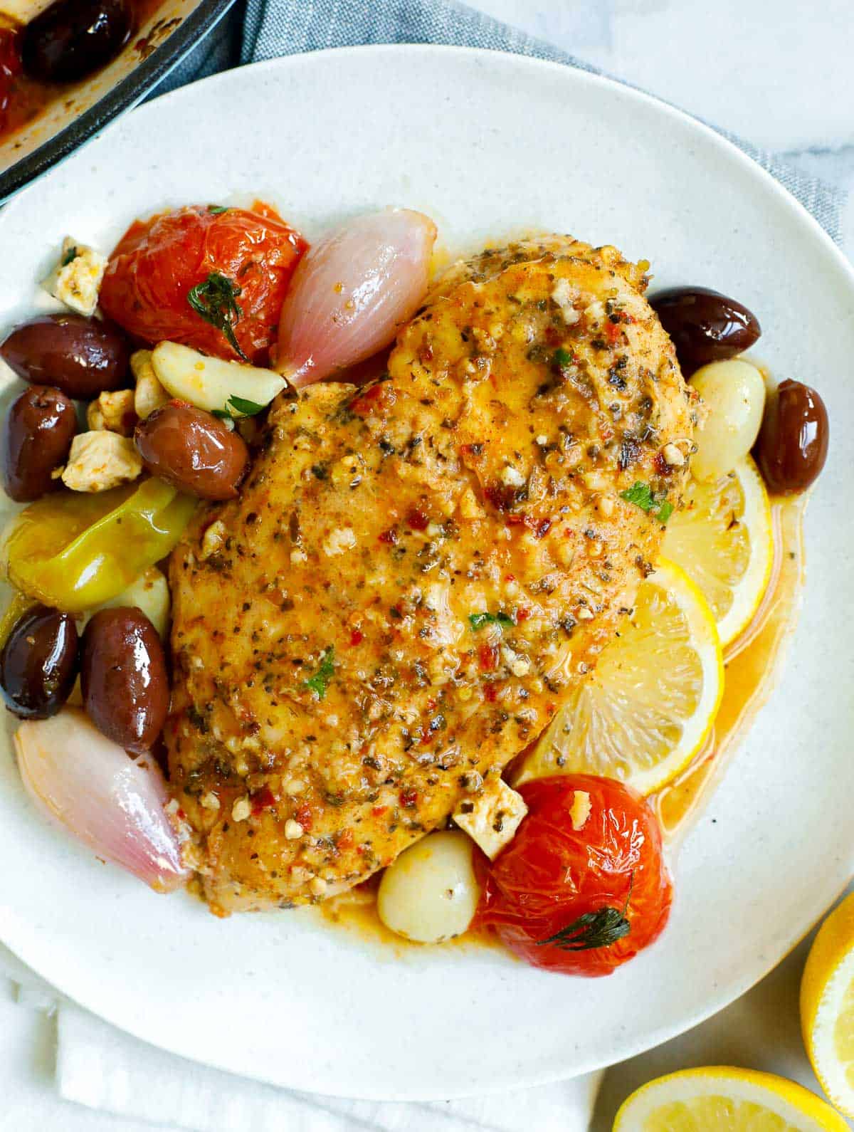 A plate with a chicken breast, olives, shallots, lemon slices, and tomatoes.