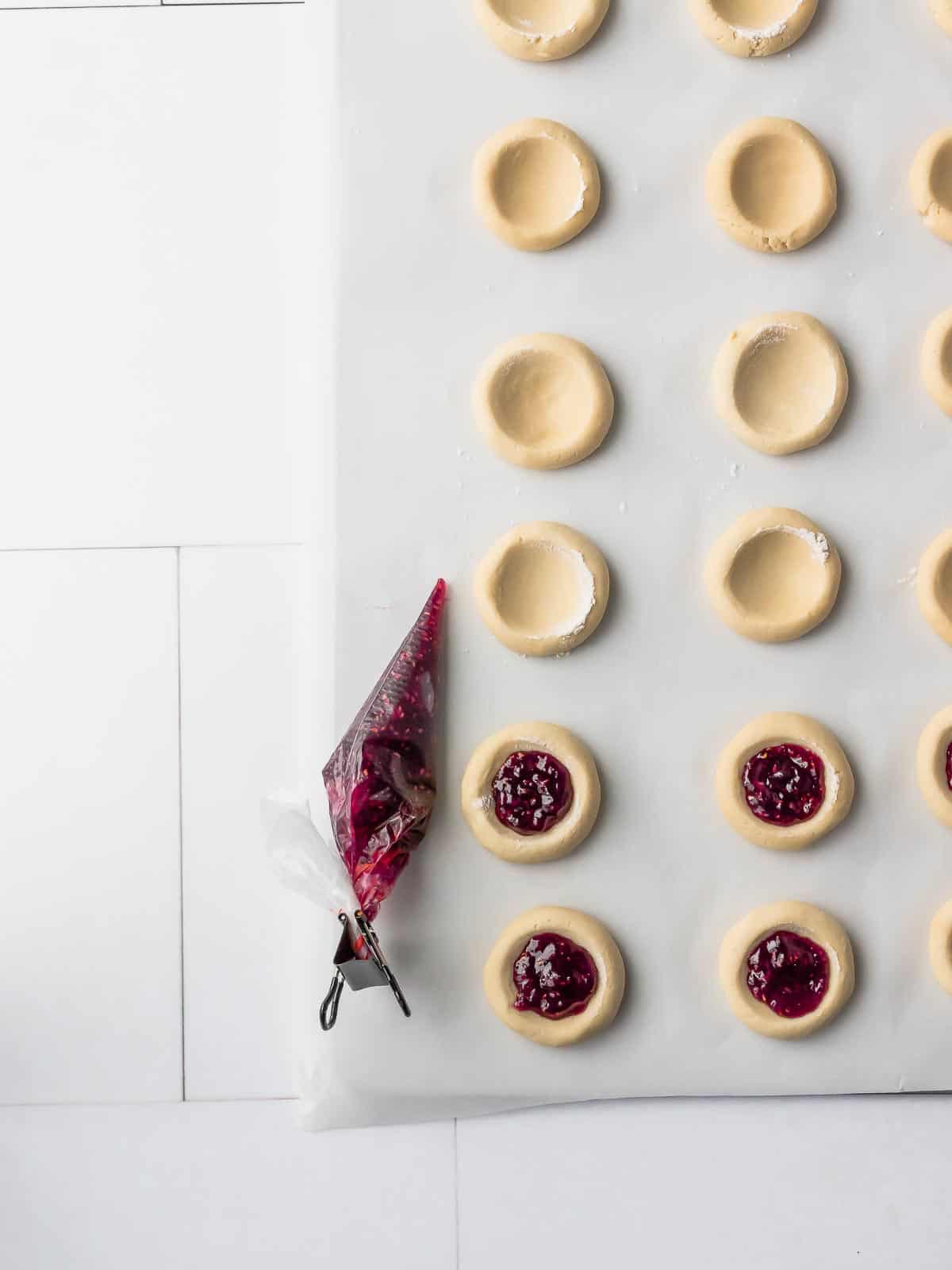 Raspberry jam piped into the center indents of cookie dough on a lined baking sheet.