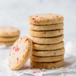 Buttery shortbread cookies with red peppermint pieces stacked on parchment paper.