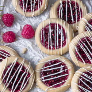 Raspberry-filled cookies topped with thin white chocolate lines beside fresh raspberries.