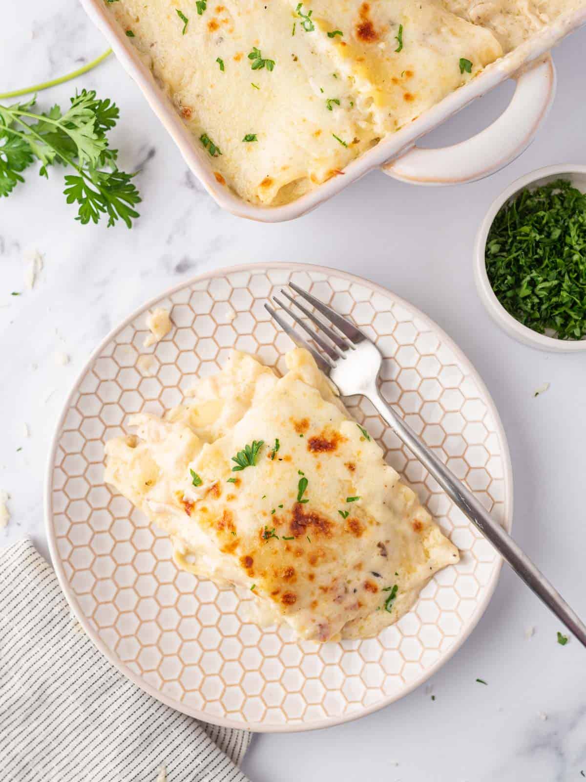 White chicken lasagna on a white plate with a silver fork and the remaining casserole in the background.