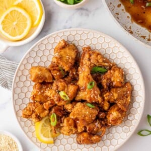 A white plate with Asian Lemon Chicken and a fresh lemon wedge. A bowl of fresh lemons is off to the side.