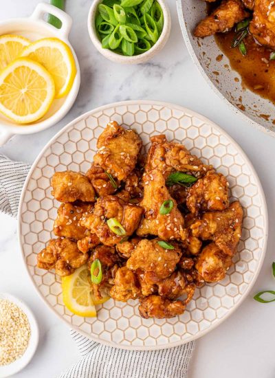 A white plate with Asian Lemon Chicken and a fresh lemon wedge. A bowl of fresh lemons is off to the side.