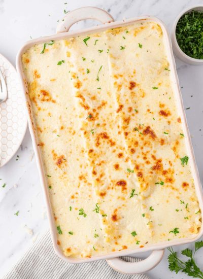 White chicken lasagna in a casserole dish with a plate and a silver fork to the side and a bowl of chopped parsley.