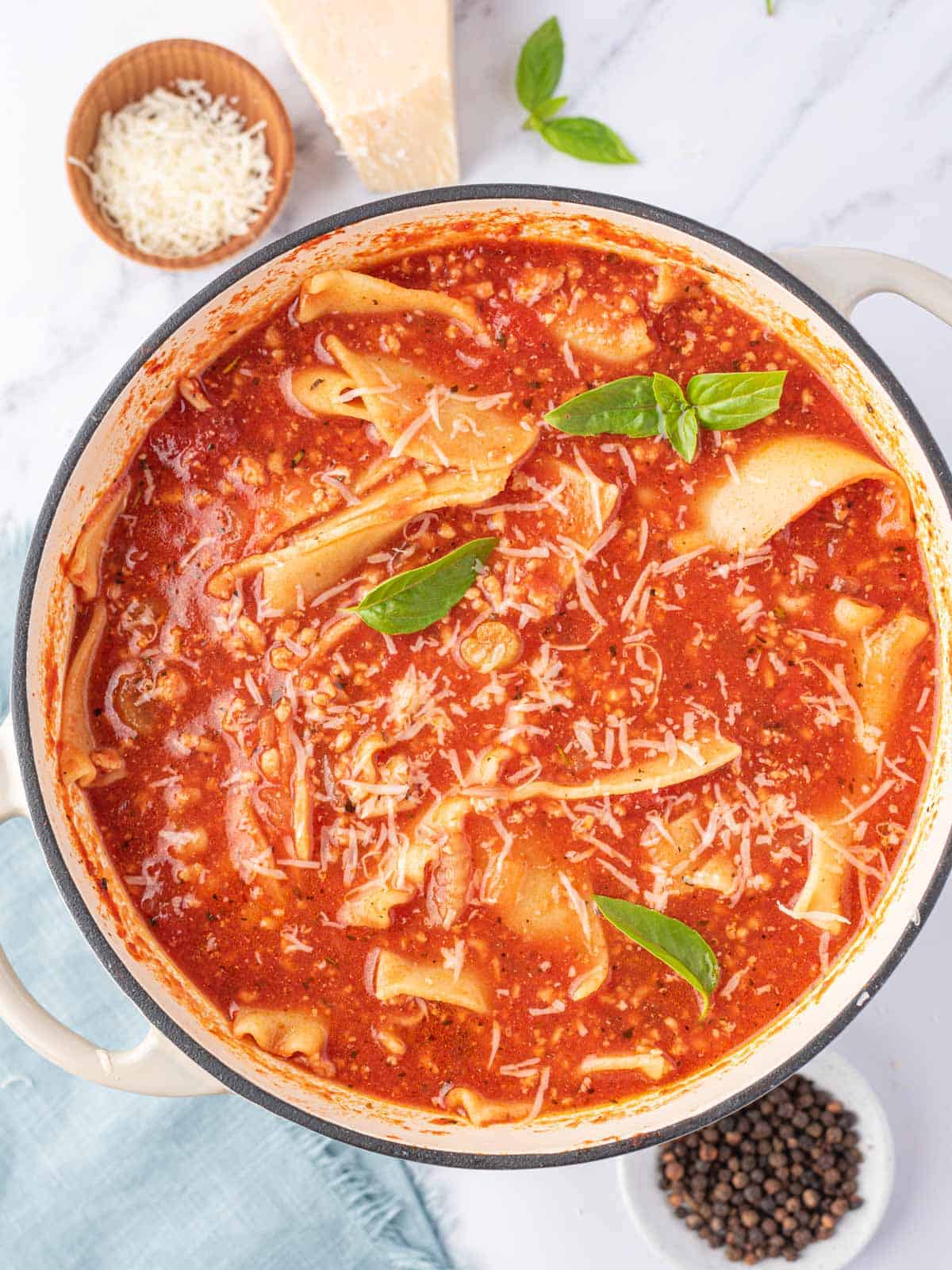 A top view of one pot lasagna soup with a few leaves of green basil and a small bowl of parmesan cheese to the side.