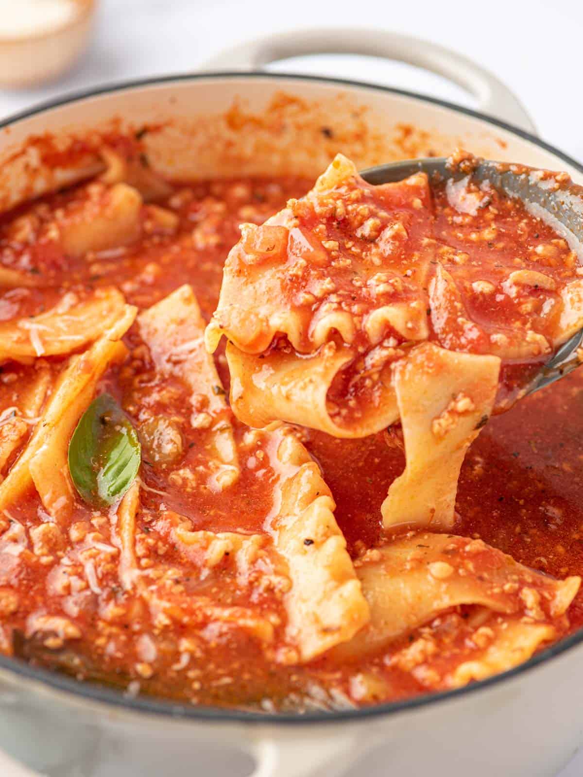 A ladle is being used to scoop noodle and tomato broth out of a white enameled dutch oven filled with easy lasagna soup.