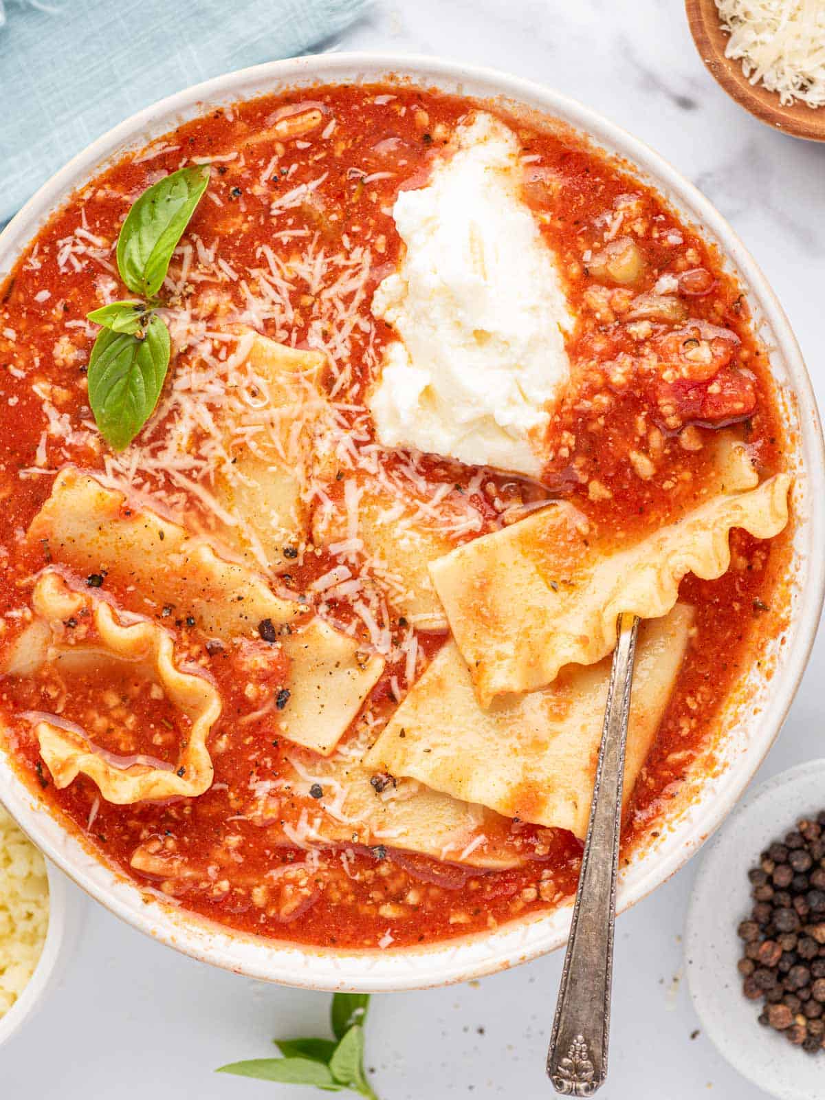 A silver spoon in a bowl of easy lasagna soup with basil leaves, parmesan cheese and a dollop of ricotta cheese.