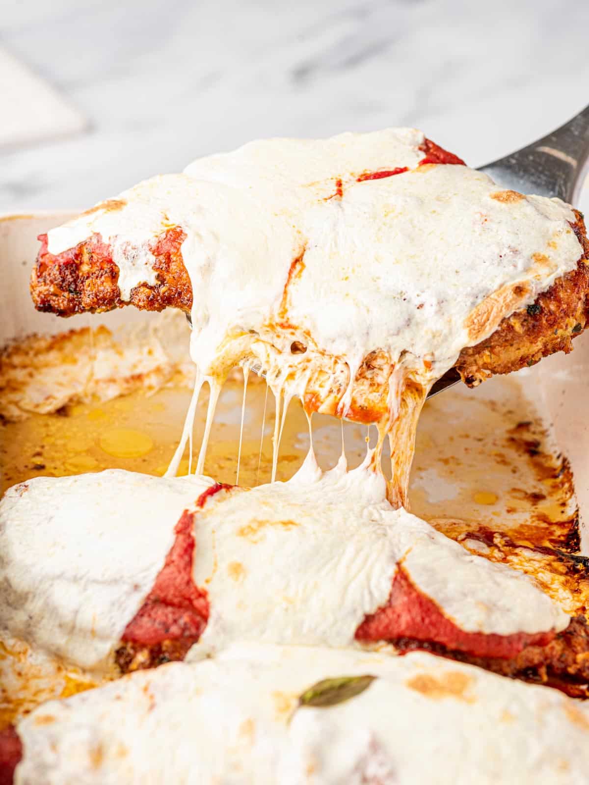 A spatula lifts a piece of easy baked chicken parm out of a baking dish. Stringy cheese is stretching from the dish.