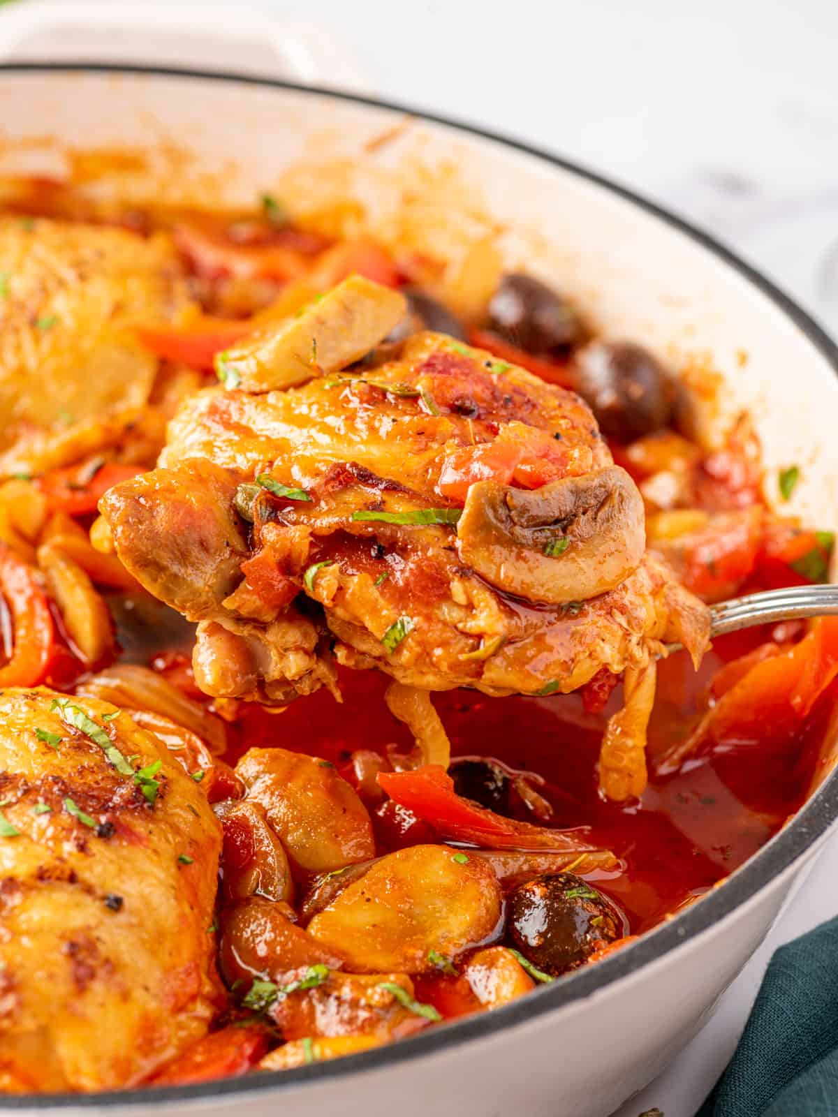 A closeup photo of a skillet of chicken with a chicken thigh on a silver spoon. cacciatore