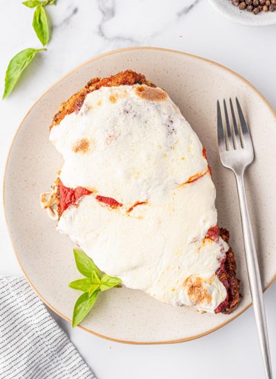 Oven Baked Chicken Parmesan is on an off-white plate with a silver fork and a few leaves of fresh basil.