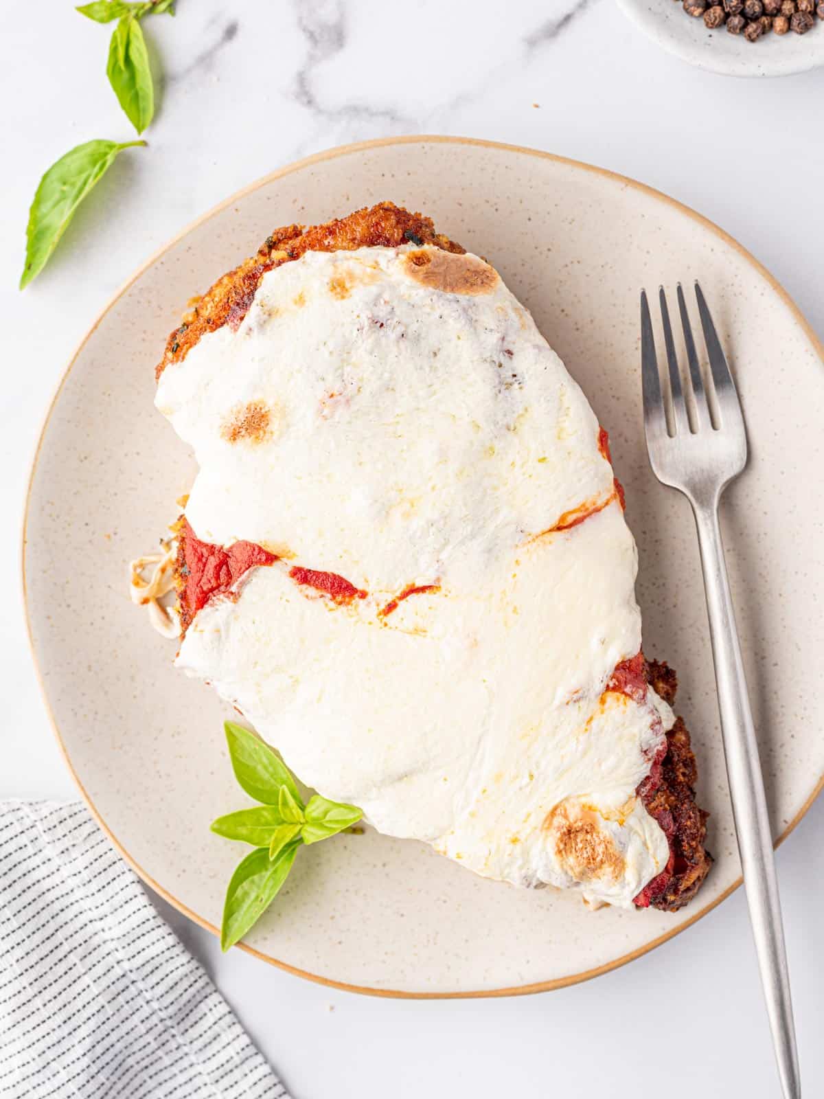 Oven Baked Chicken Parmesan is on an off-white plate with a silver fork and a few leaves of fresh basil.