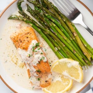 A plate with a fillet of salmon with creamy lemon butter sauce with asparagus and lemon slices.