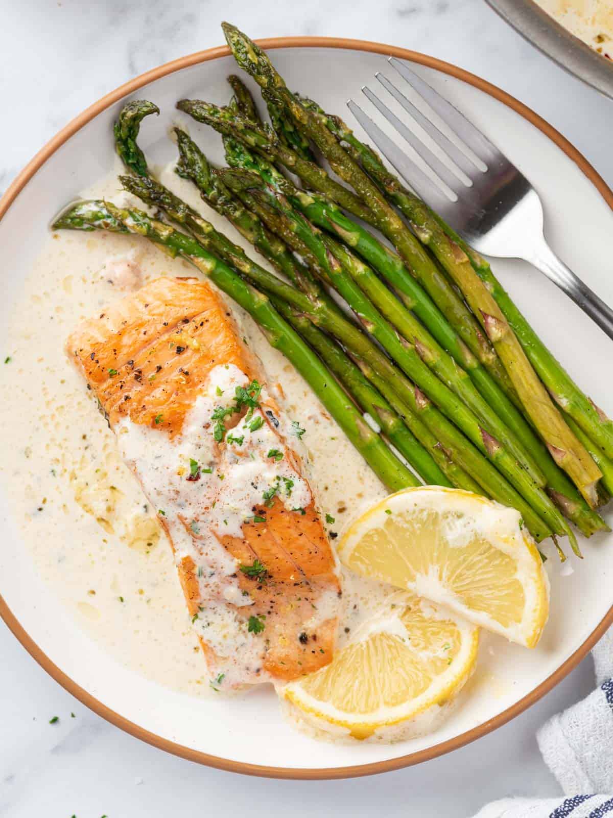 A plate with a fillet of salmon with creamy lemon butter sauce with asparagus and lemon slices.