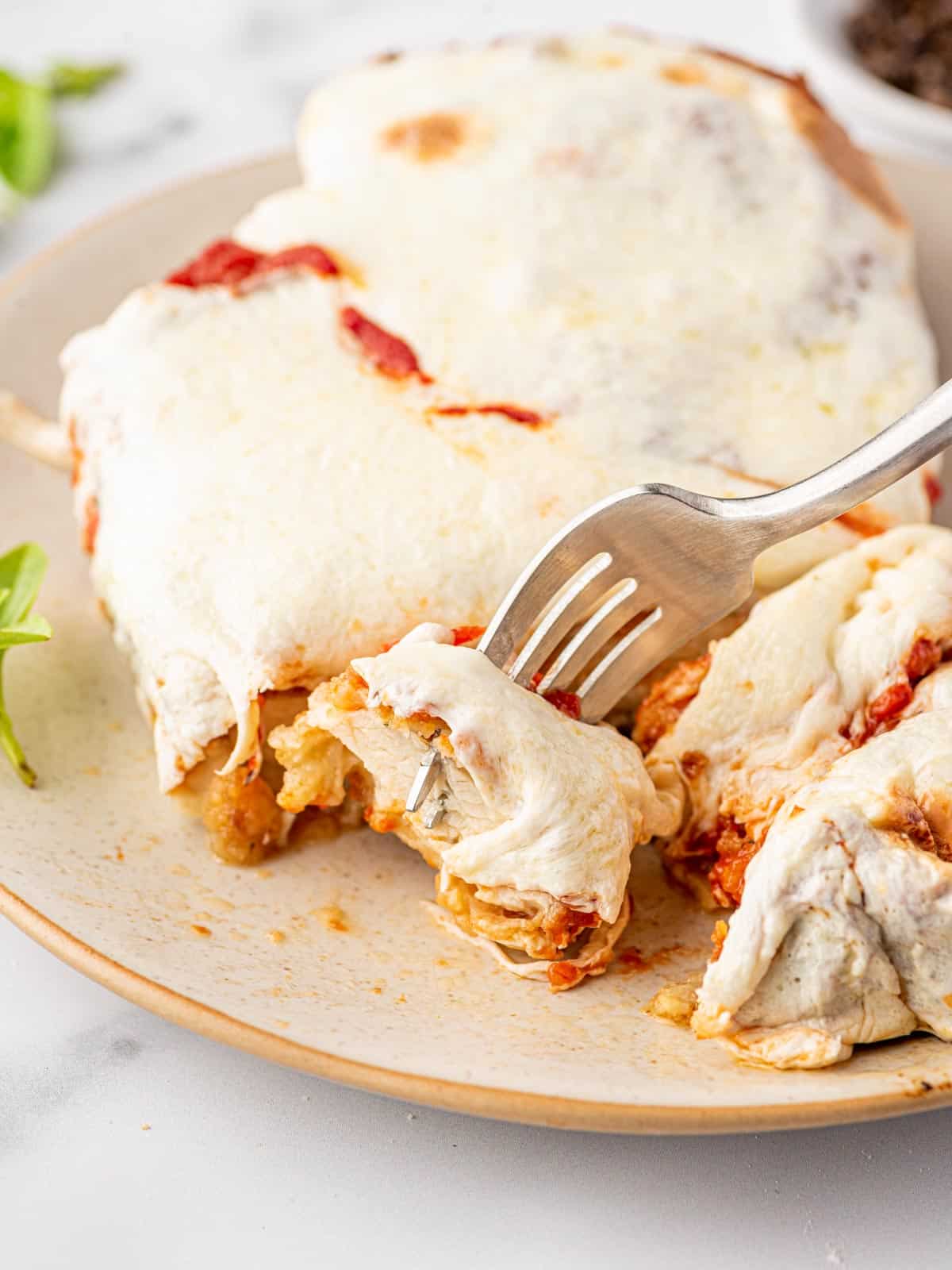 A silver fork is holding a bite of parmesan crusted chicken topped with marinara and mozzarella. The rest of the chicken is on the plate.