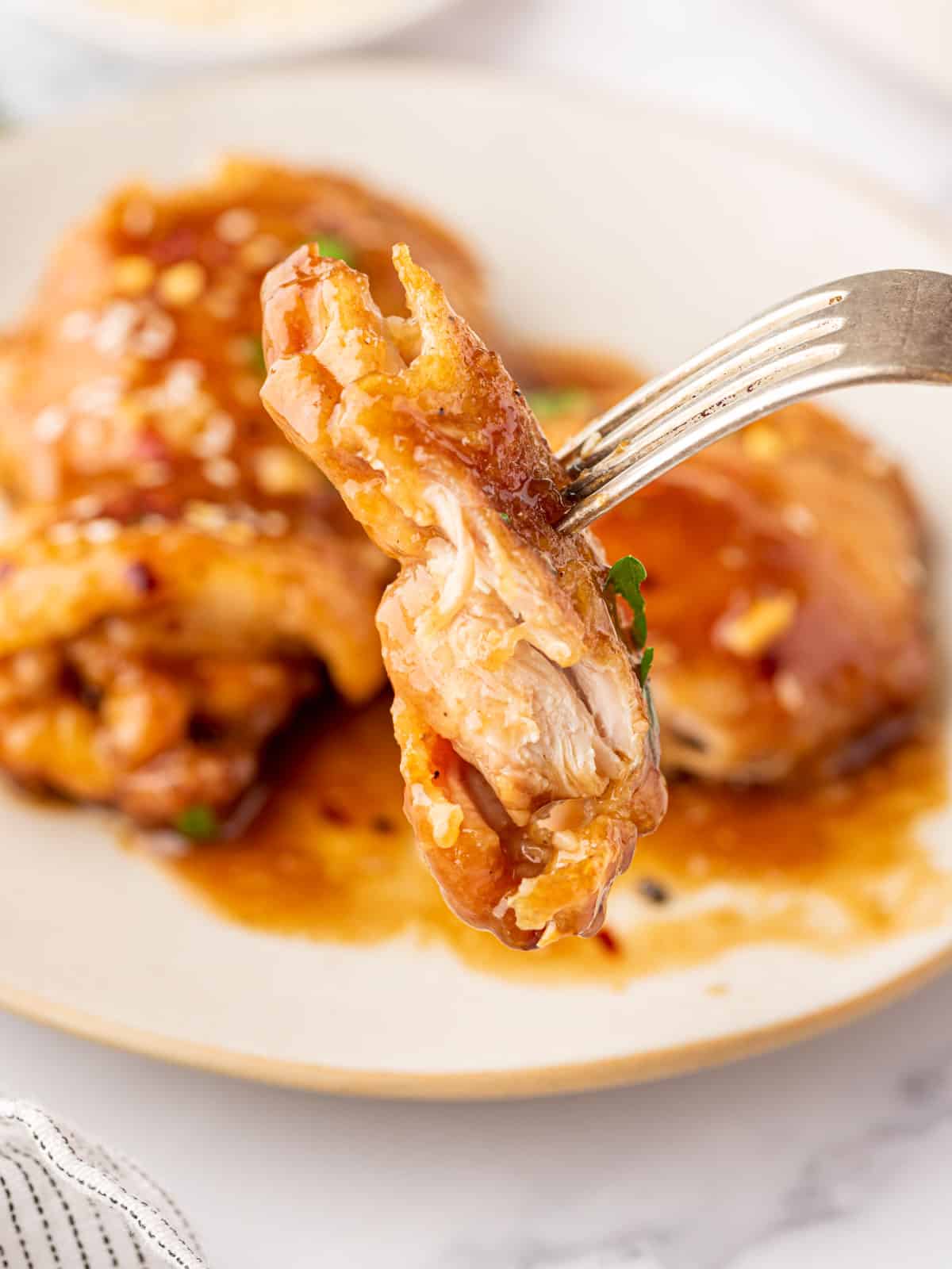 Juicy chicken thigh meat on a fork with a plate of honey garlic chicken in the background.