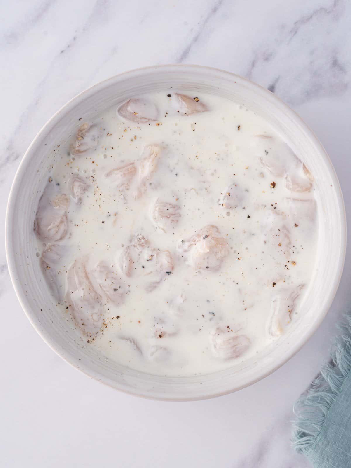 Raw chicken nuggets marinating in a bowl of seasoned buttermilk.