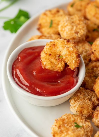 A piece of air fryer popcorn shrimp is laying in a bowl of cocktail sauce.