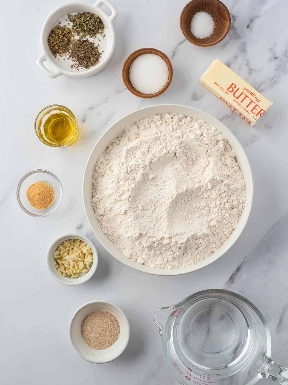 Ingredients for garlic knots arranged with flour, yeast, butter, garlic, herbs, olive oil, and water.