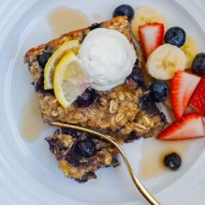 A square of blueberry oatmeal bake on a plate topped with whipped cream and fresh fruit.