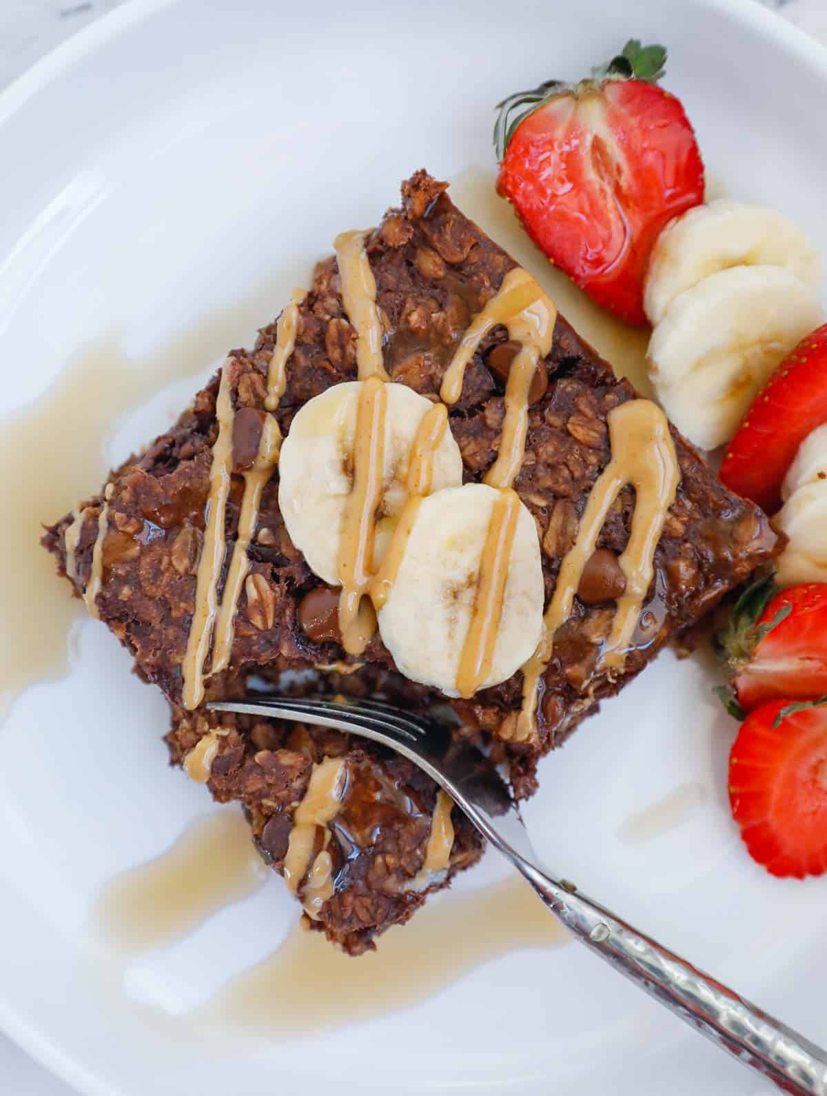piece of baked chocolate oats on a plate, topped with slices of bananas and drizzle of peanut butter, as well as maple syrup.