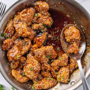A spoon with sesame coated fried chicken rests near the edge of a pan of honey sesame chicken.