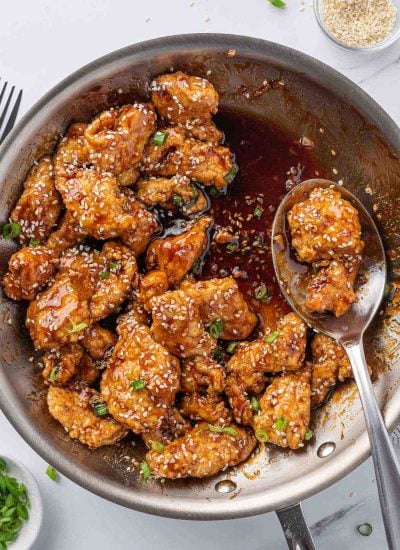 A spoon with sesame coated fried chicken rests near the edge of a pan of honey sesame chicken.