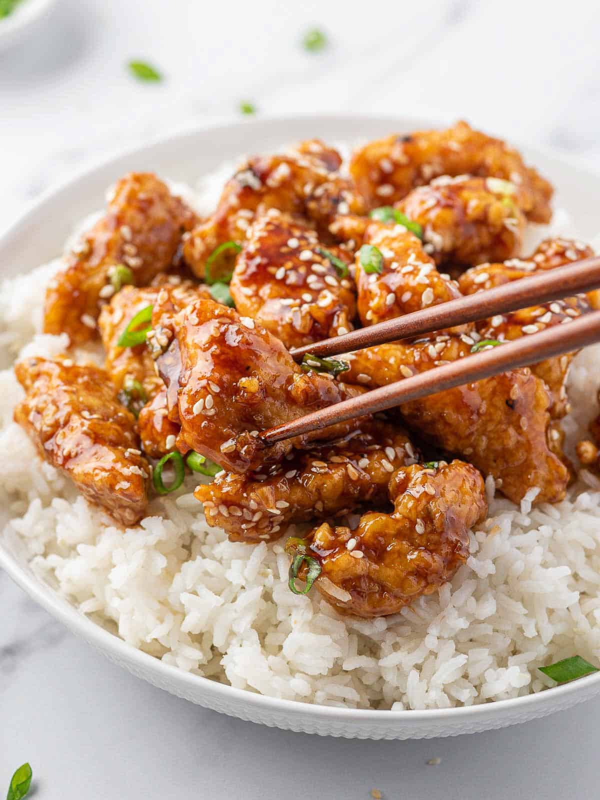 Chopstick hold a piece of crispy honey sesame chicken on a bed of white rice.