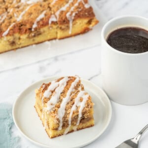 A slice of keto coffee cake on a white plate next to a cup of coffee and a fork. A tray of almond flour coffee cake sits in the background.