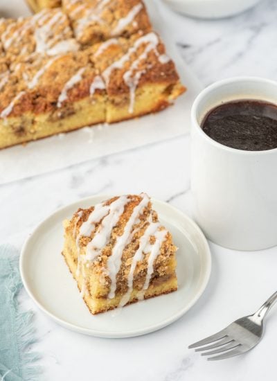 A slice of keto coffee cake on a white plate next to a cup of coffee and a fork. A tray of almond flour coffee cake sits in the background.