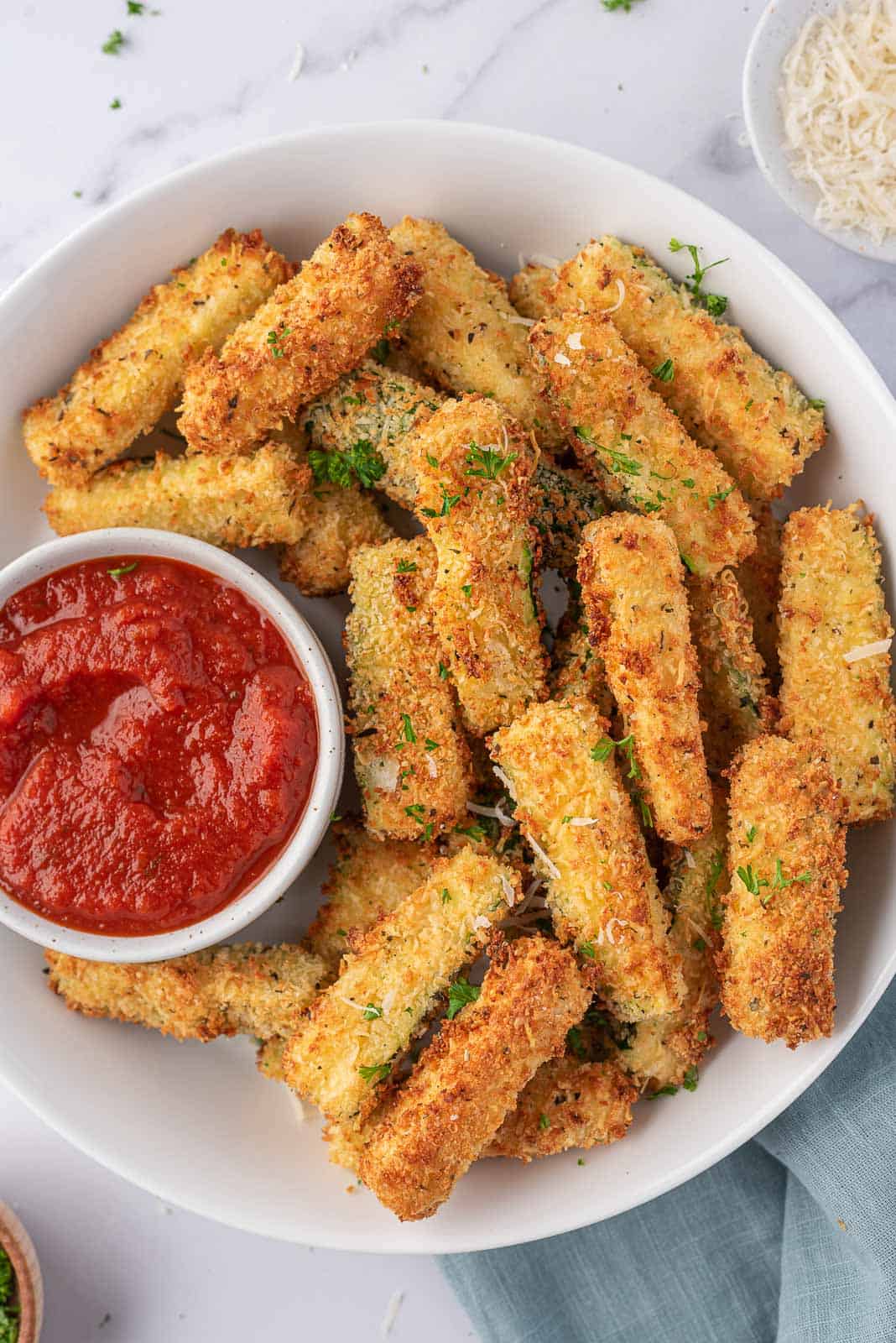 Air fryer zucchini sticks on a plate with a bowl of marinara.