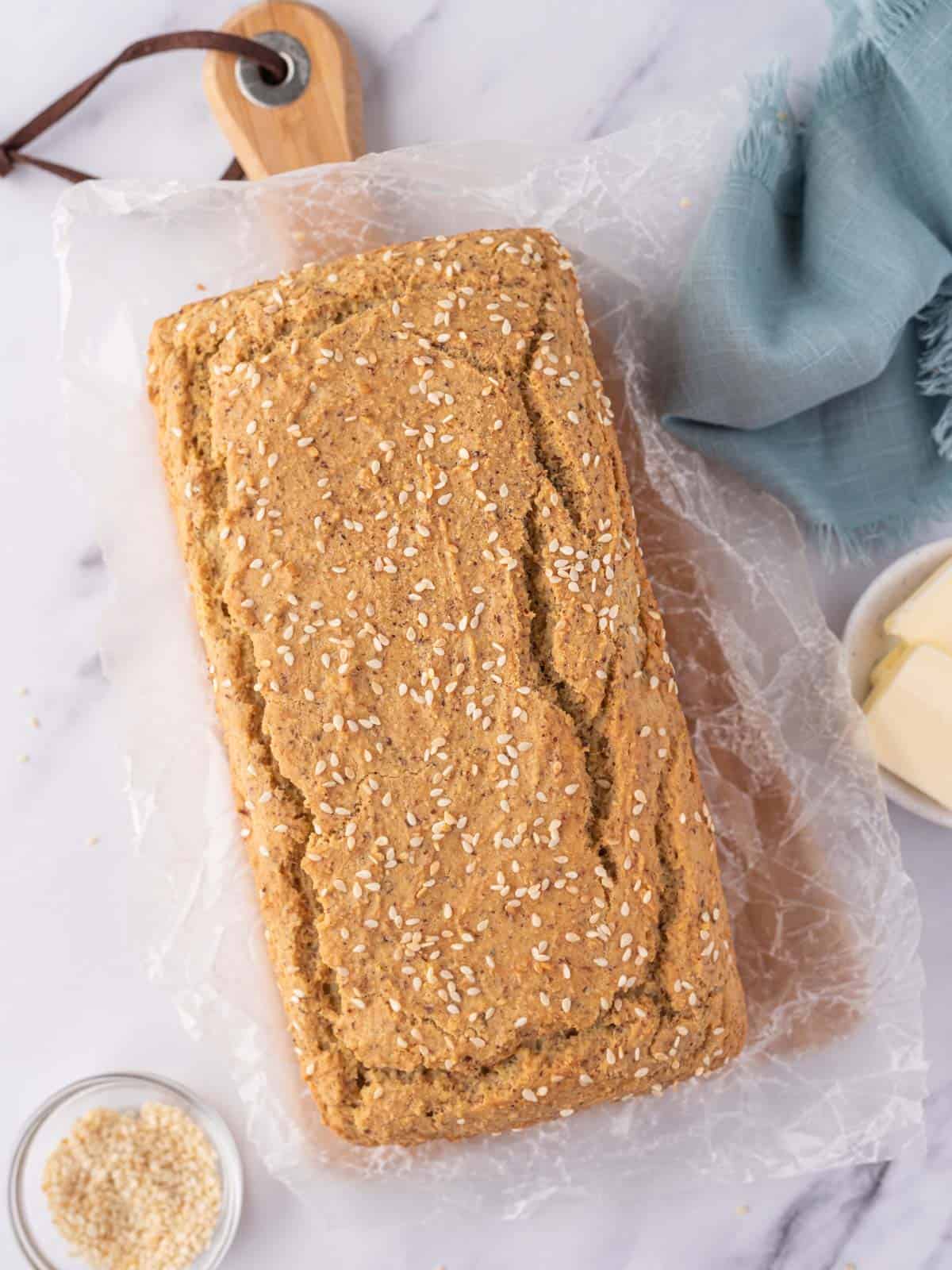 A loaf of low carb sandwich bread topped with sesame seeds rests on a wax paper covered bread board.