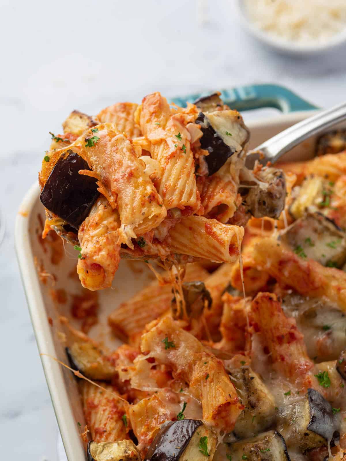A scoop of baked eggplant pasta with the casserole dish in the background.