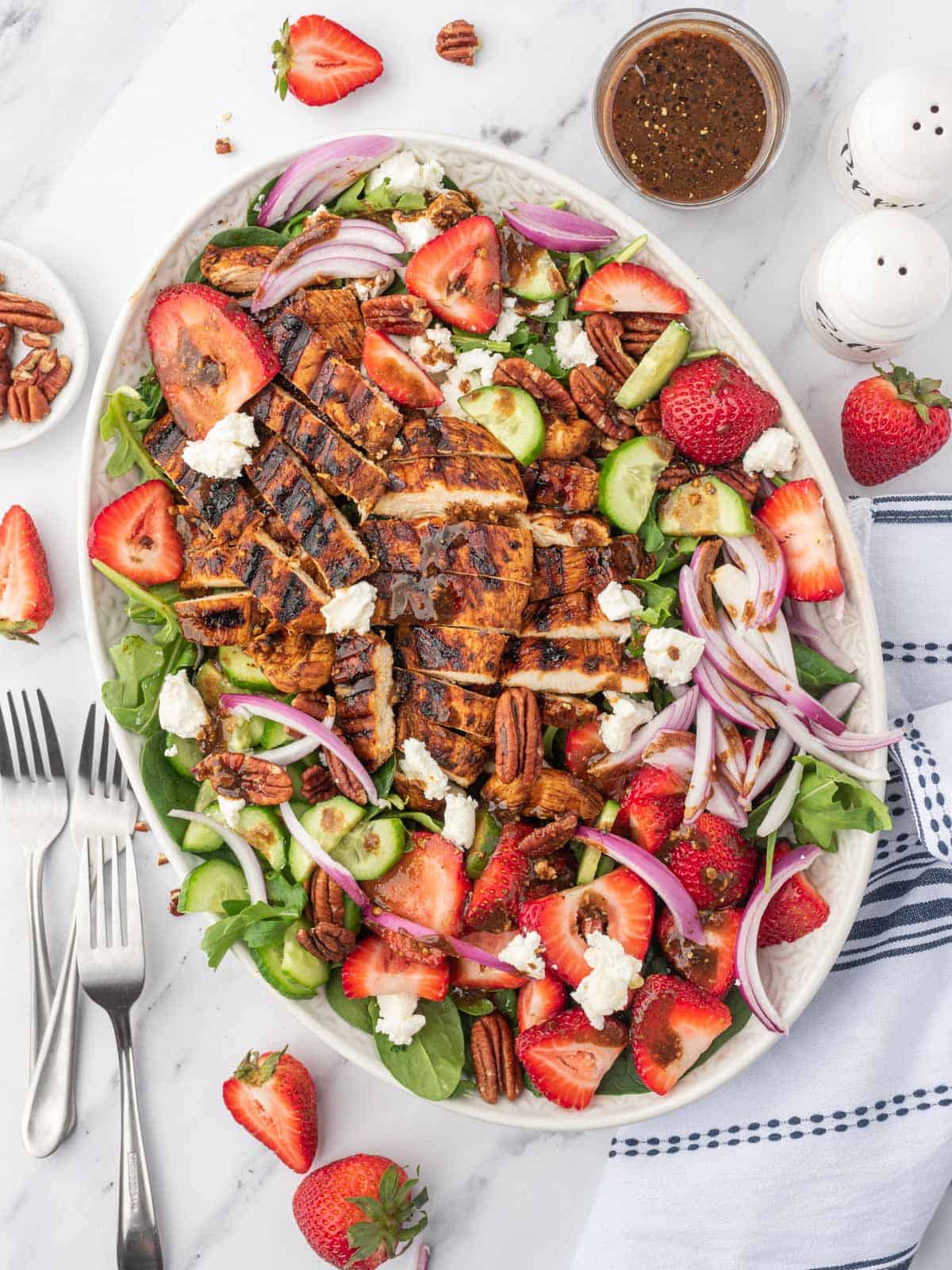 A large platter of spinach salad with strawberries and balsamic grilled chicken with several forks on the side.