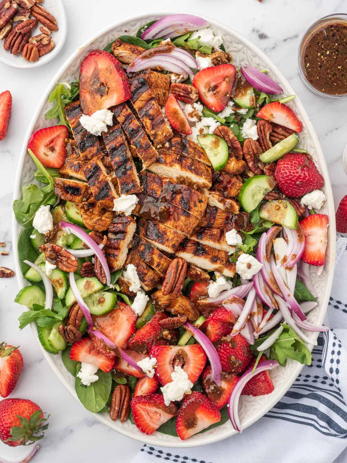 Grilled chicken on spinach salad with fresh strawberries on a serving platter with balsamic dressing and pecans to the side.