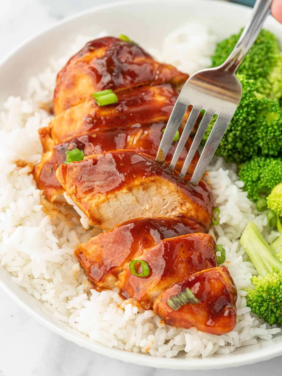 A fork picks up a piece of sliced bbq chicken breast from a plate of rice.