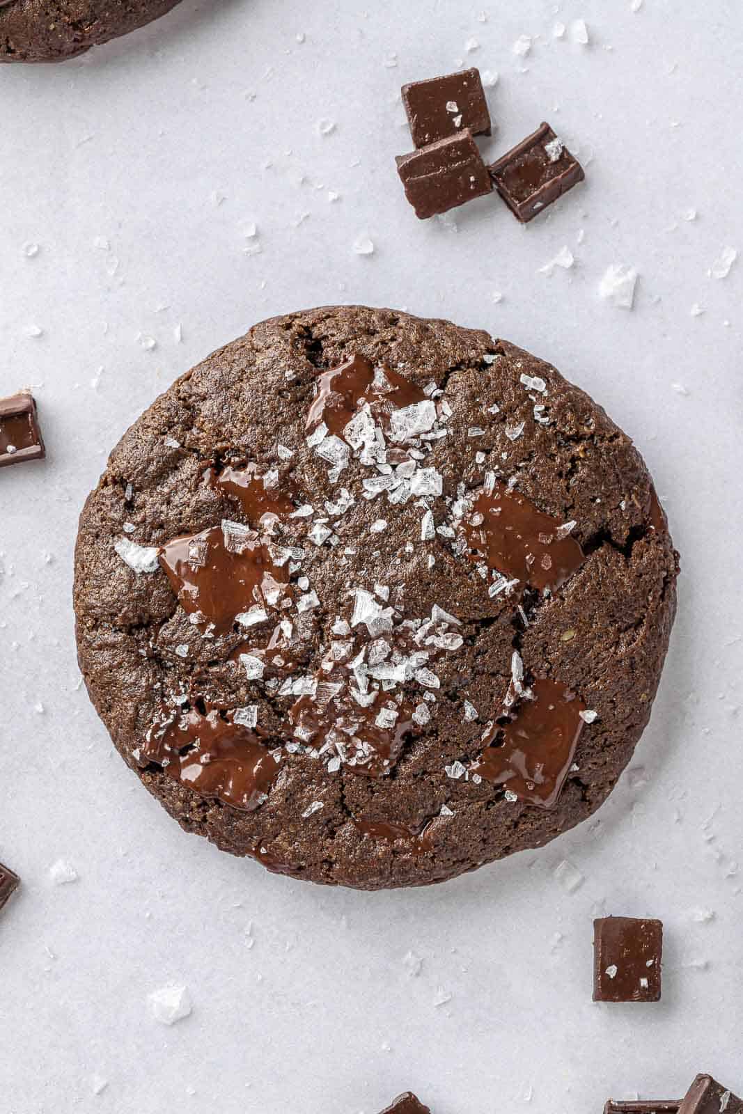 A single vegan double chocolate cookie on a white surface with chocolate chunks and salt.