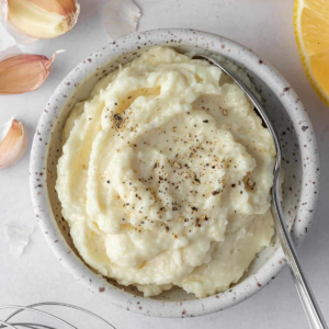 Thick, creamy Lebanese garlic sauce in a speckled ceramic bowl with a spoon, garlic cloves, and lemon wedges nearby.