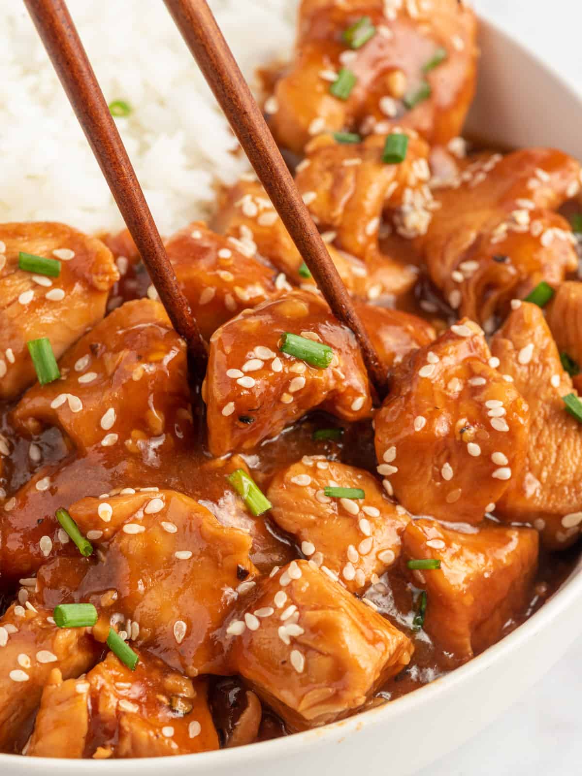 Chopsticks pick up a piece of sesame chicken from a bowl.