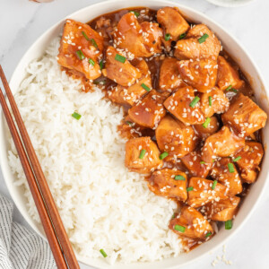 Chinese sesame chicken in a bowl with rice and chopsticks resting on the side.