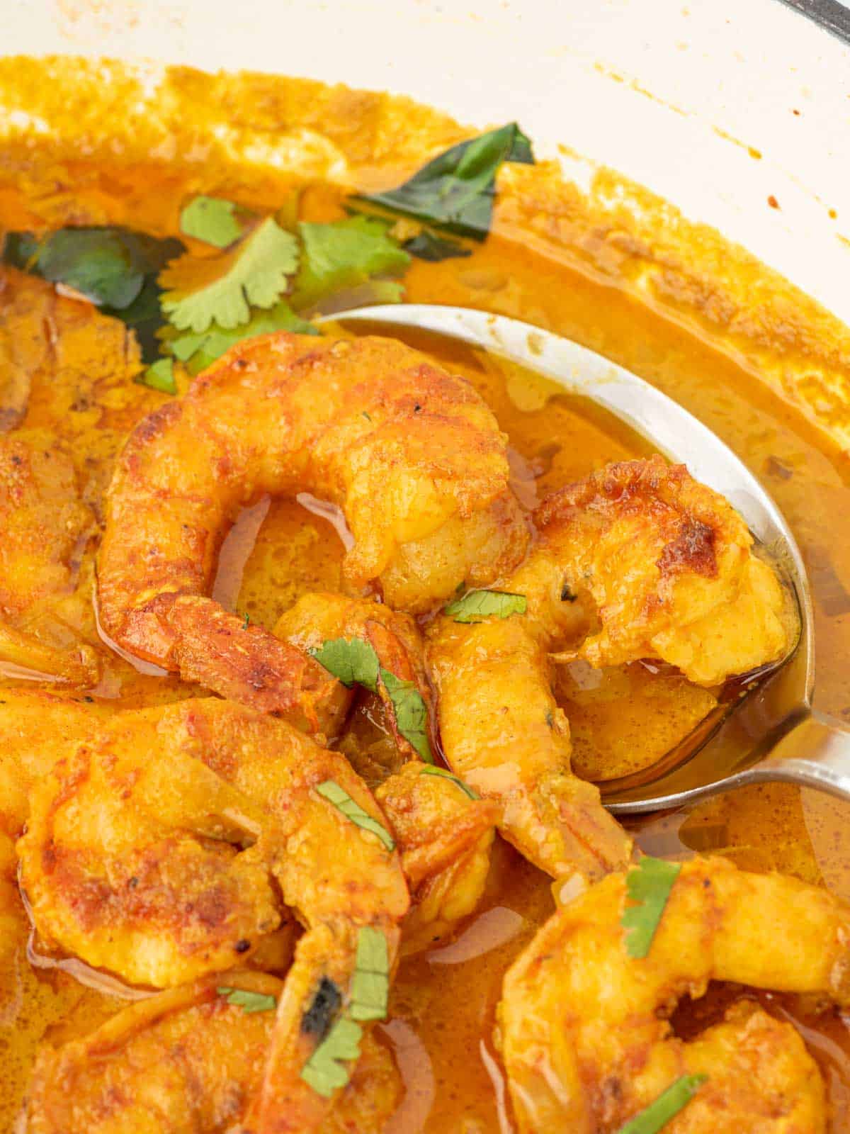 A spoon lifts two pieces of shrimp from a pot of red curry sauce.