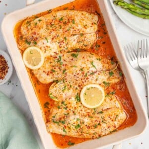 A baking dish with garlic butter tilapia filets and lemon garnish.