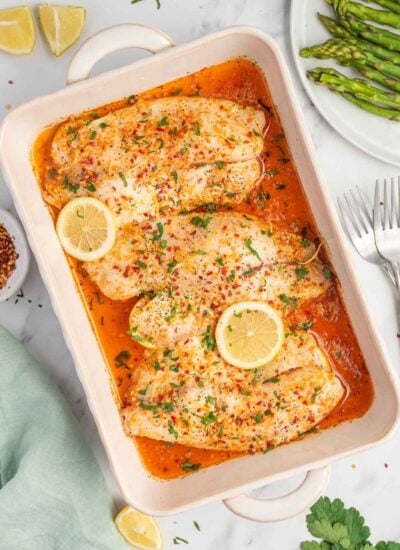 A baking dish with garlic butter tilapia filets and lemon garnish.