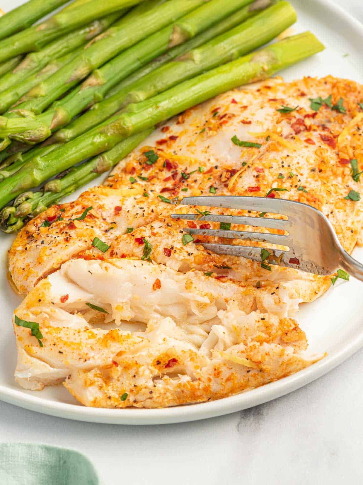 A fork flakes a garlic butter tilapia filet on a plate.