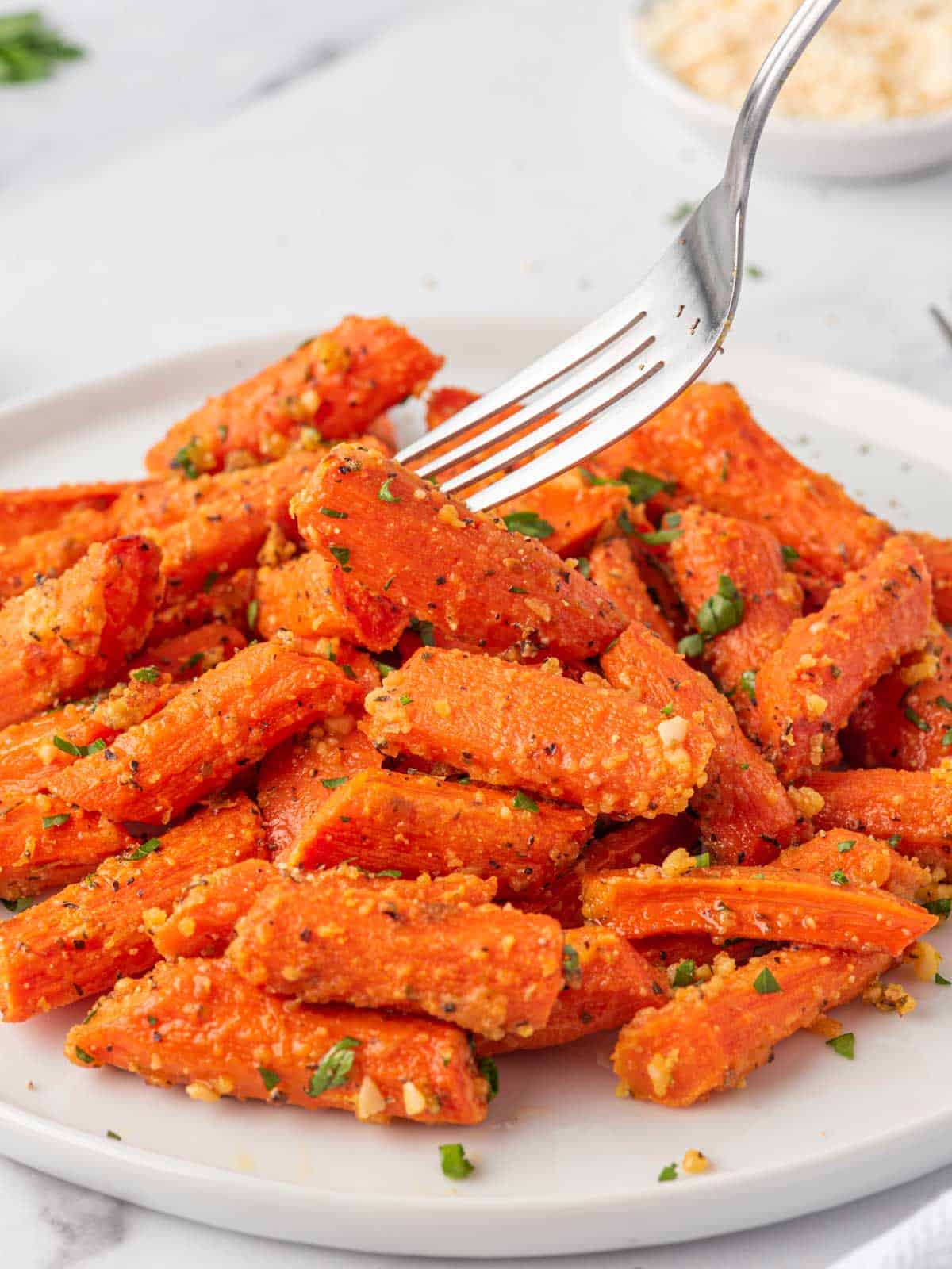 Fork lifting a Parmesan roasted carrot from a plate garnished with chopped parsley.