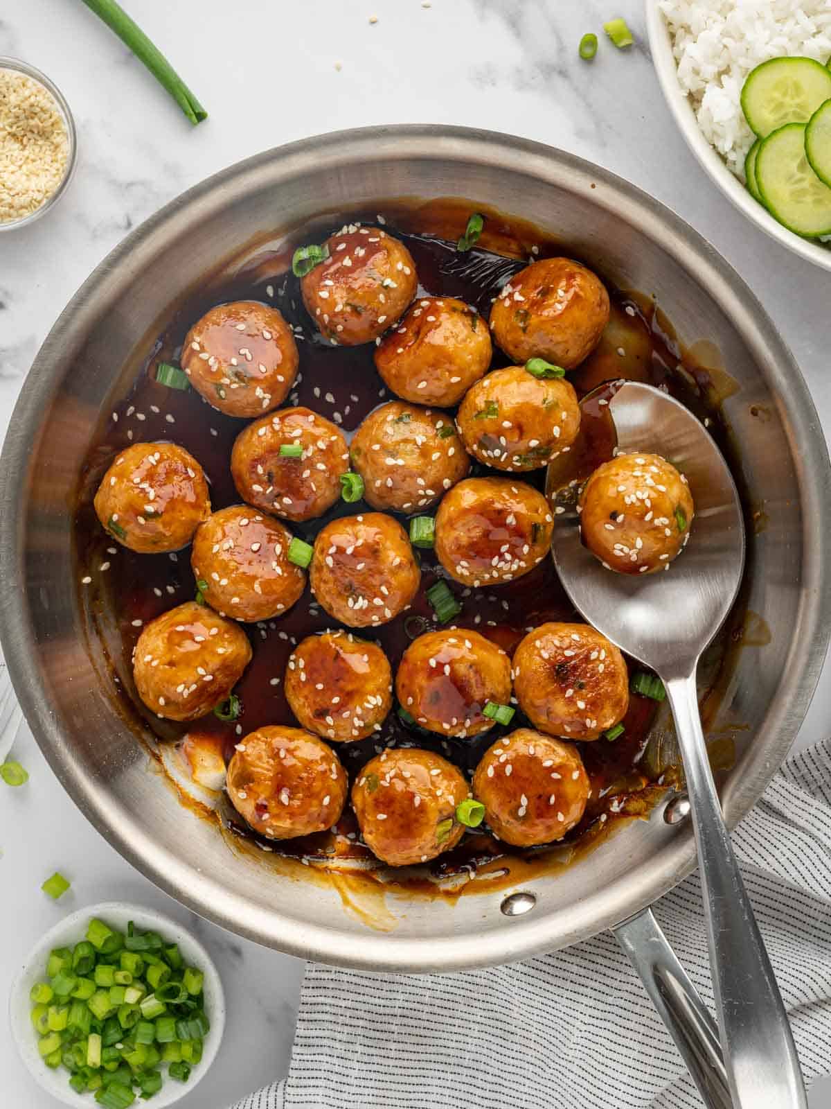 A spoon lifts a teriyaki meatball from a skillet.
